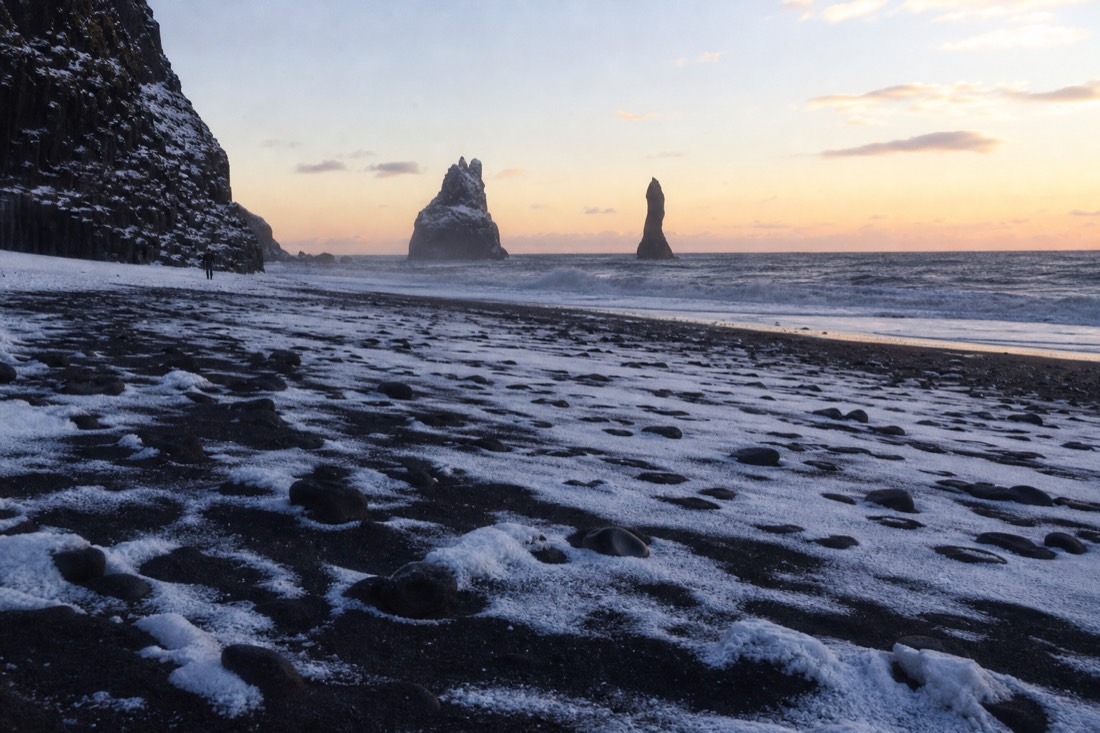 Snow lying on black sand in low winter sun, Reynisdrangar sea stacks offshore, calm after the storm