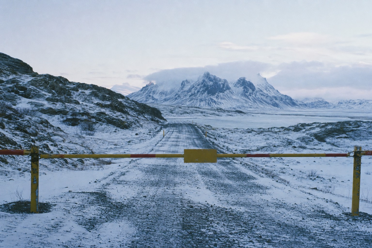 Empty snow-covered gravel road in Iceland closed by a barrier with mountains behind