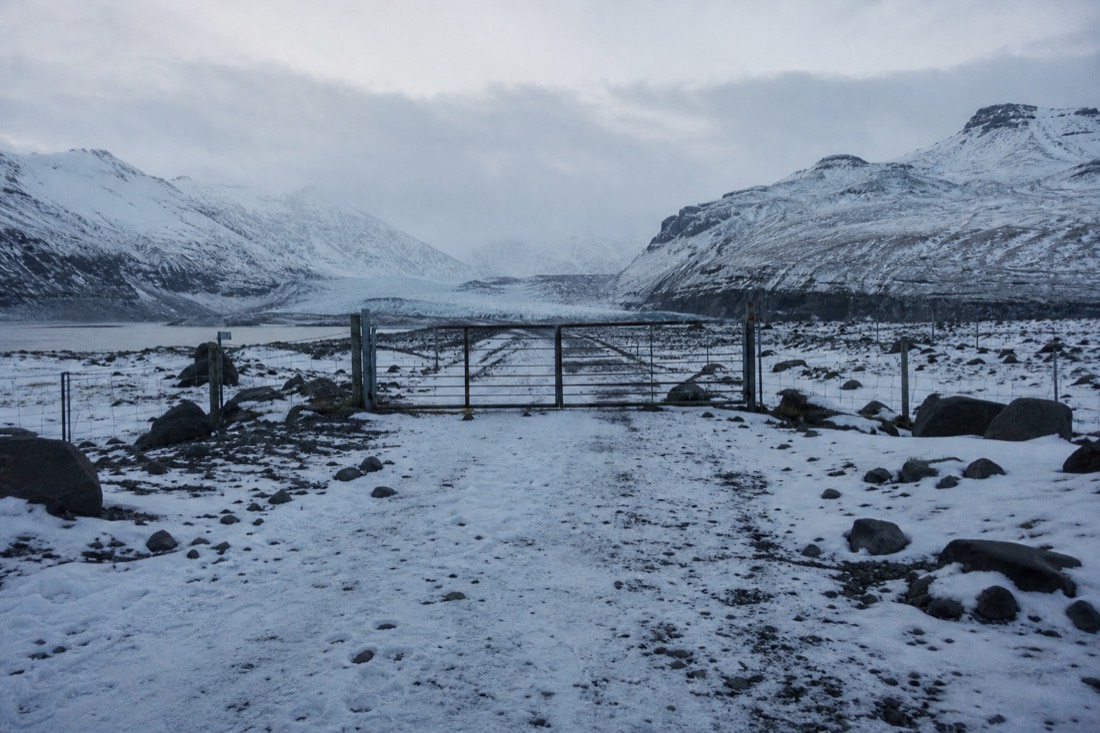 Snow-covered gravel track behind a closed metal gate, mountains beyond, flat grey January light
