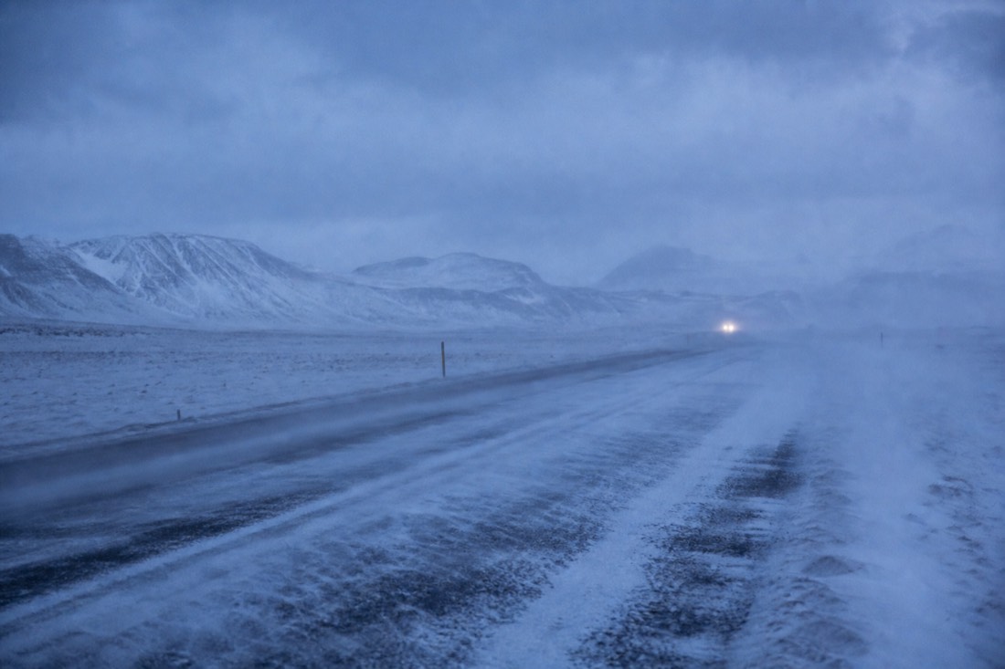 Snow blowing across an empty ring road at dusk, one distant pair of headlights, low visibility