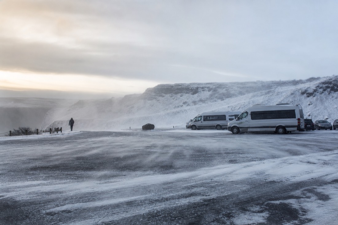 Empty snow-blown car park near Gullfoss with parked tour vehicles, wind lifting spindrift, flat grey light