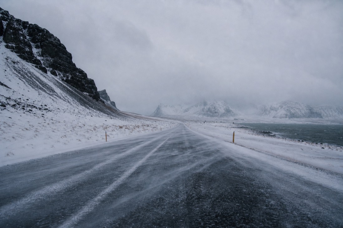 Snow drifting across an empty closed stretch of Icelandic ring road under flat grey light