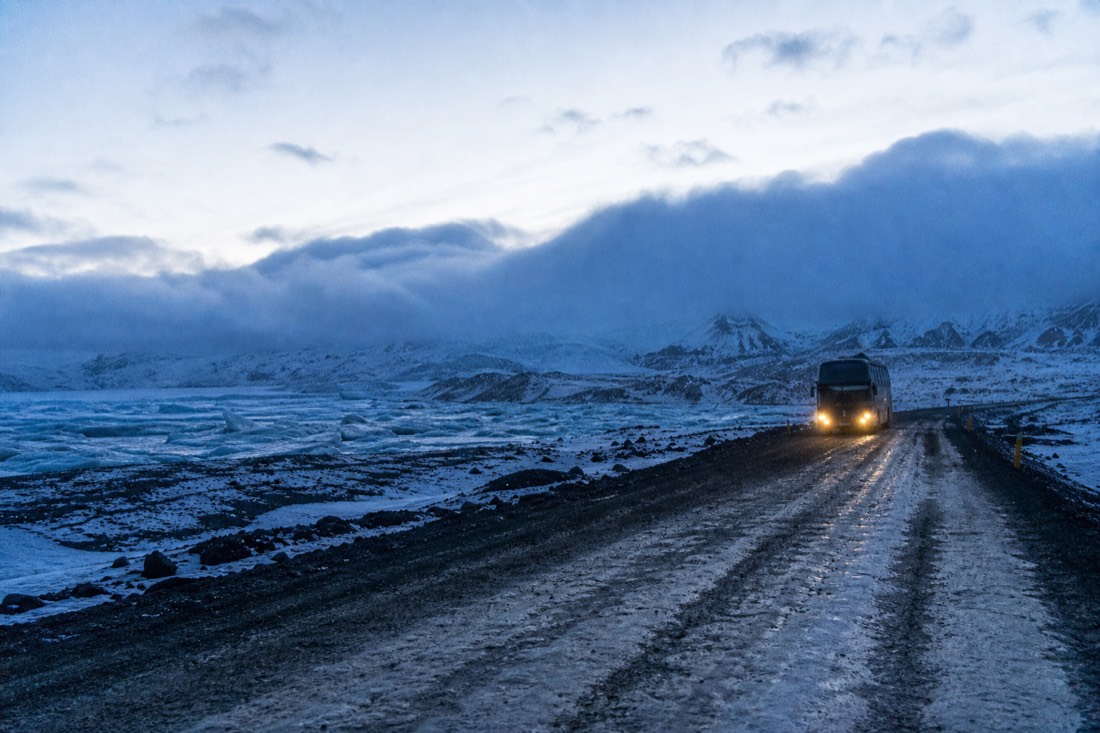 Coach headlights sweeping a snowy gravel track as it turns away from a bank of low cloud