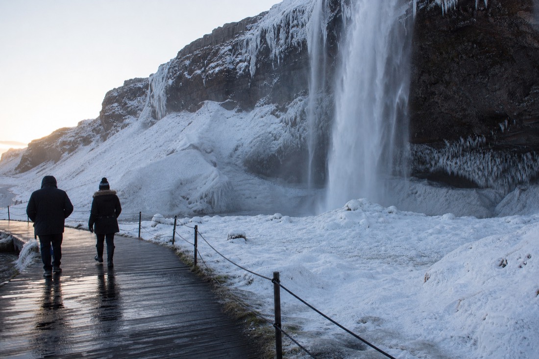 Half-frozen Seljalandsfoss waterfall under pale winter light with a wet timber boardwalk below