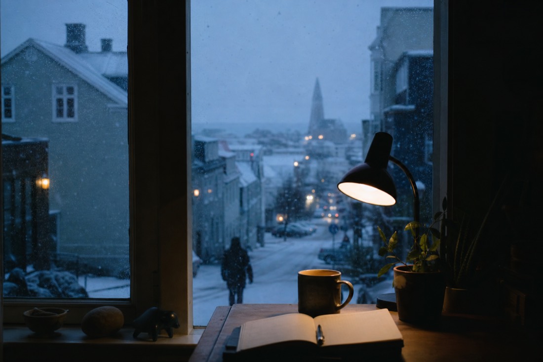 Lamplit desk by a window on a dark Reykjavik afternoon, notebook and mug, snow falling outside
