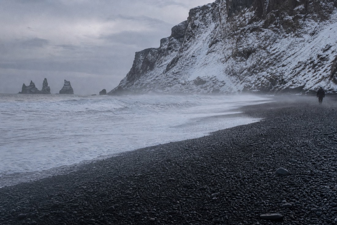 Waves washing over dark wet gravel at Reynisfjara with spray blowing inland under flat grey light
