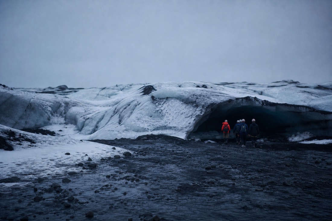 Distant helmeted figures at a cave entrance on the glacier under a flat grey sky